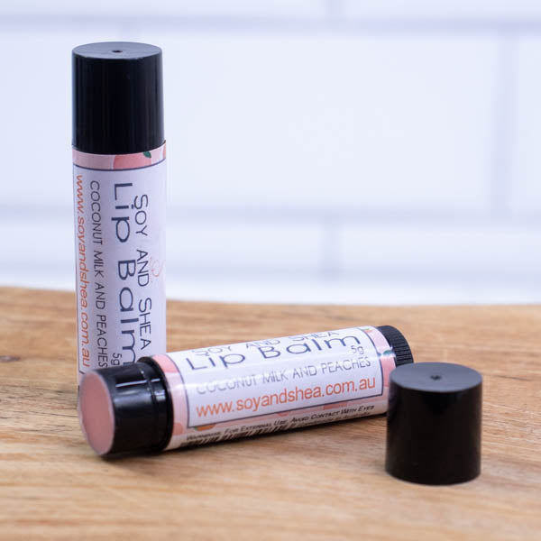 Two black tubes of lip balm are displayed on a wooden surface with blurred tiled background.  One tube stands on its end while the other lays flat in front with the cap removed revealing a peach balm.  The label is light pink with watercolour illustrations of peaches