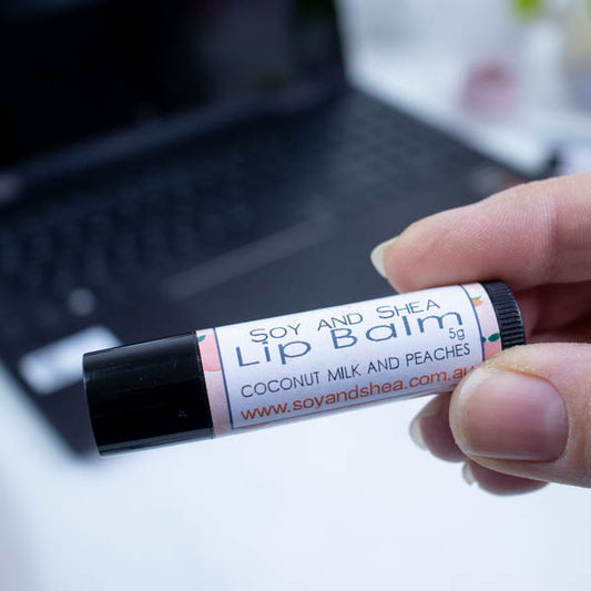 A hand holds a black tube of lip balm against a blurred background with an open laptop computer on a white surface.  The label has text LIP BALM COCONUT MILK AND PEACHES on white background with a pale orange border with drawings of peaches.