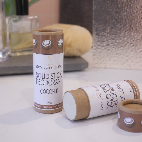 Two cardboard tubes labelled with SOLID STICK DEODORANT rest on a bathroom counter top with a blurred background.  One tube stands upright on the bench while another lays flat with the lid removed to reveal an off-white balm inside.  The label on the lid is chocolate brown with coconut halfs