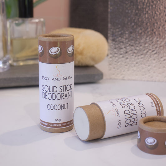 Two cardboard tubes labelled with SOLID STICK DEODORANT rest on a bathroom counter top with a blurred background.  One tube stands upright on the bench while another lays flat with the lid removed to reveal an off-white balm inside.  The label on the lid is chocolate brown with coconut halfs