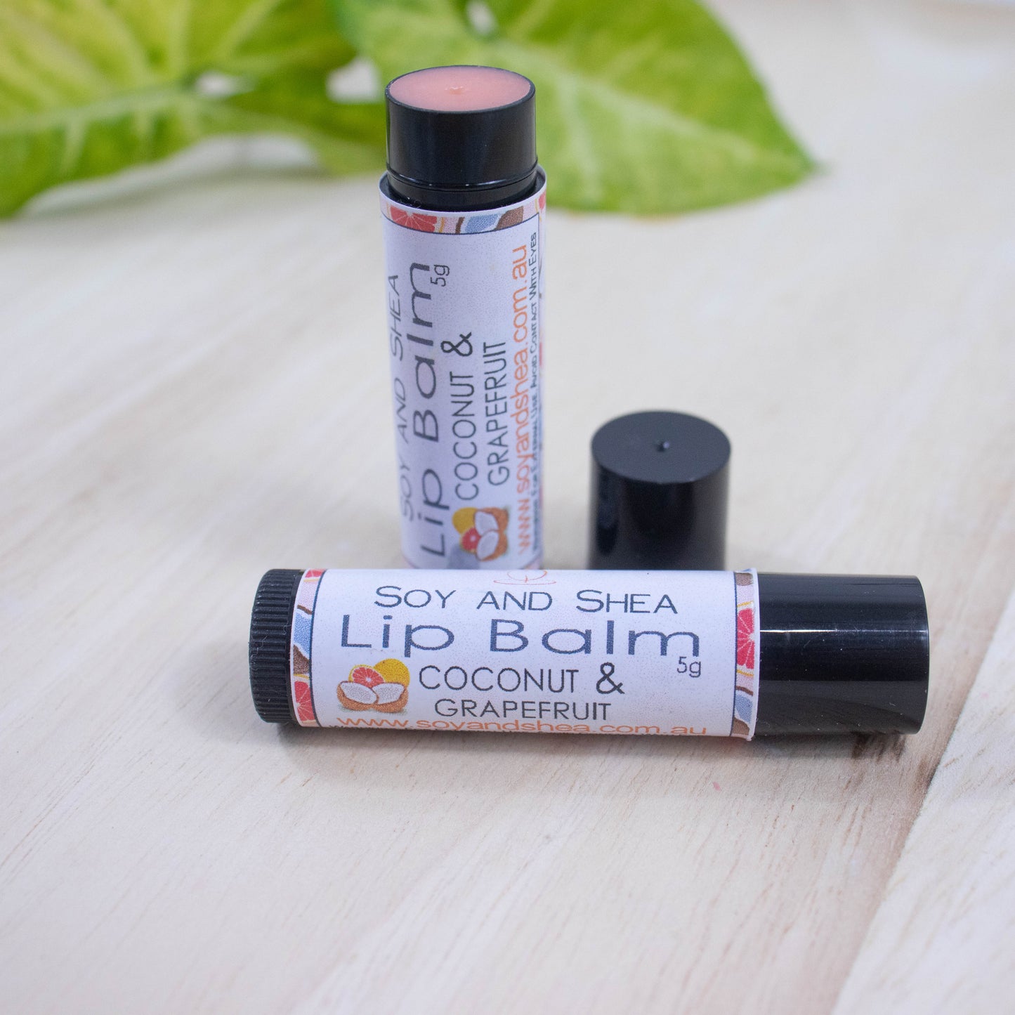 Two black tubes of lip balm displayed on a wooden surface with greenery blurred in the background.  One tube lies flat across the surface displaying the flavour while the other tube stands upright behind it with the cap removed and placed to the side revealing a peach balm.