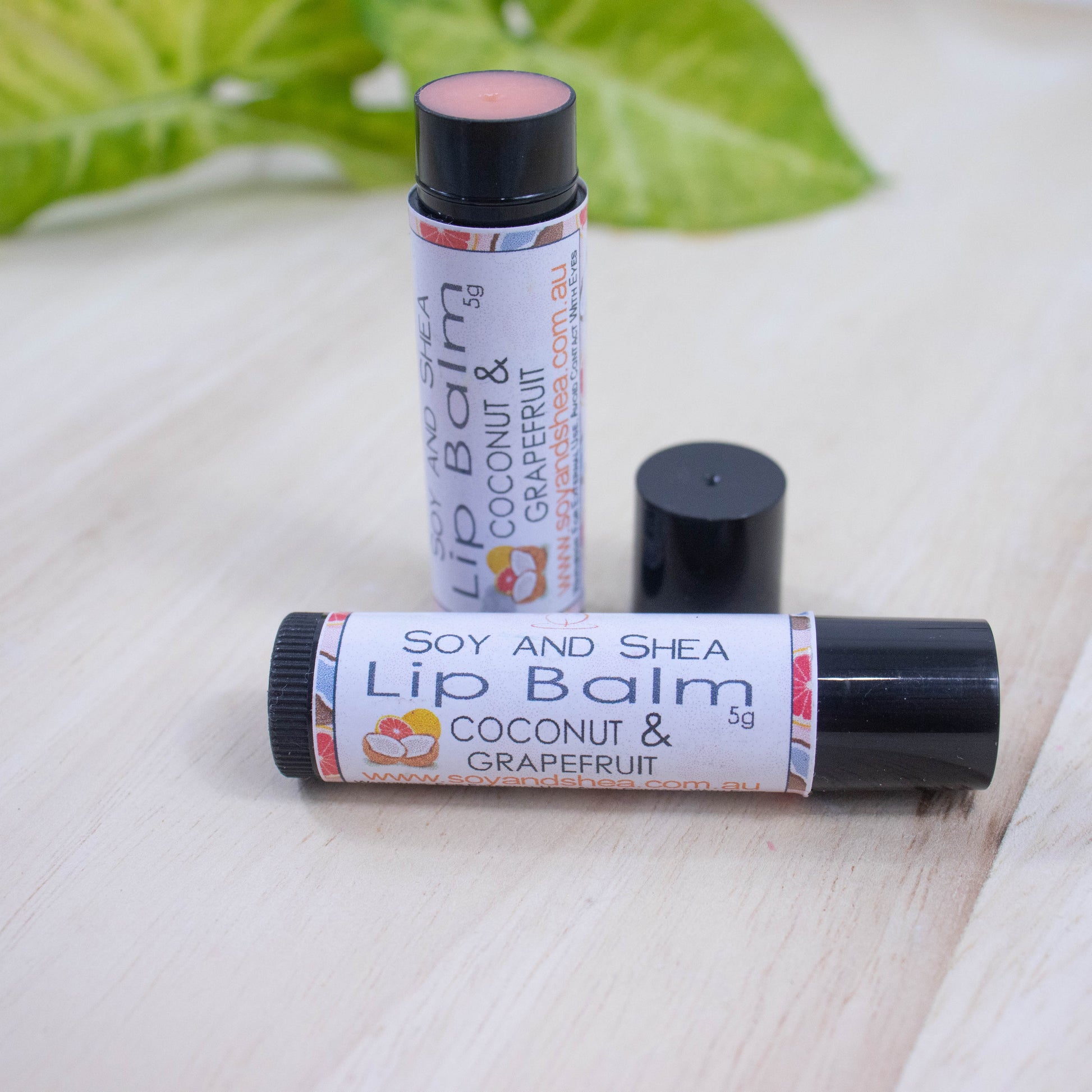 Two black tubes of lip balm displayed on a wooden surface with greenery blurred in the background.  One tube lies flat across the surface displaying the flavour while the other tube stands upright behind it with the cap removed and placed to the side revealing a peach balm.