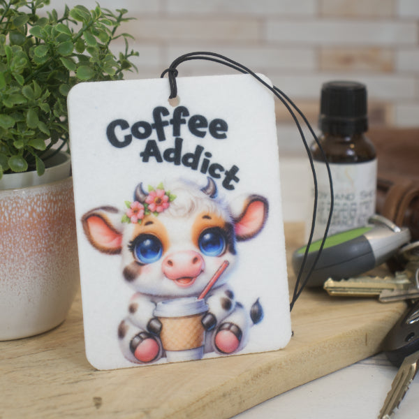 A car air freshener with a cartoon baby cow holding a coffee with straw and the words COFFEE ADDICT rests against a flower pot next to a bottle of scent and a bunch of keys
