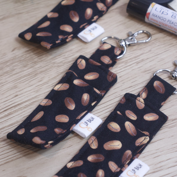 A selection of three lip balms showing variations in fabric design, lie on a wooden side  with a lip balm lays near-by.  The Clip balms have a randomly scattered brown coffee beans scattered over a black background