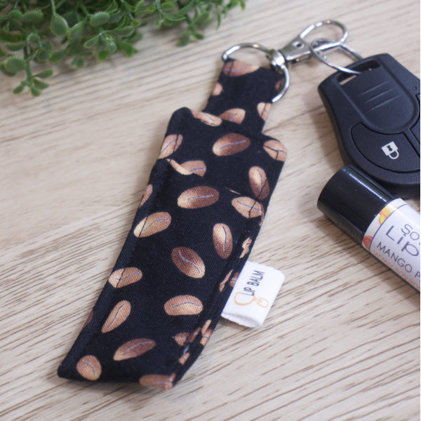 A Lip Balm holder lies on an wooden surface with its lobster claw hook attached to a car key.  A lip balm and greener can be seen in the image.  The Clip Balm has simple drawings of brown coffee beans randomly placed over a black background