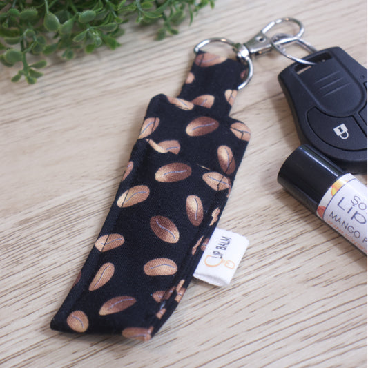 A Lip Balm holder lies on an wooden surface with its lobster claw hook attached to a car key.  A lip balm and greener can be seen in the image.  The Clip Balm has simple drawings of brown coffee beans randomly placed over a black background