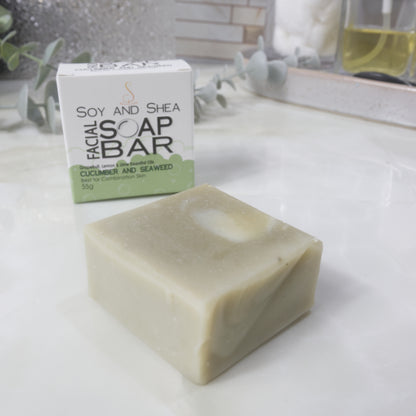A small square bar of pale green soap in the foreground with its white and light green box behind it on a light countertop. The box reads 'Soy and Shea Facial Soap Bar' and 'Cucumber and Seaweed'.