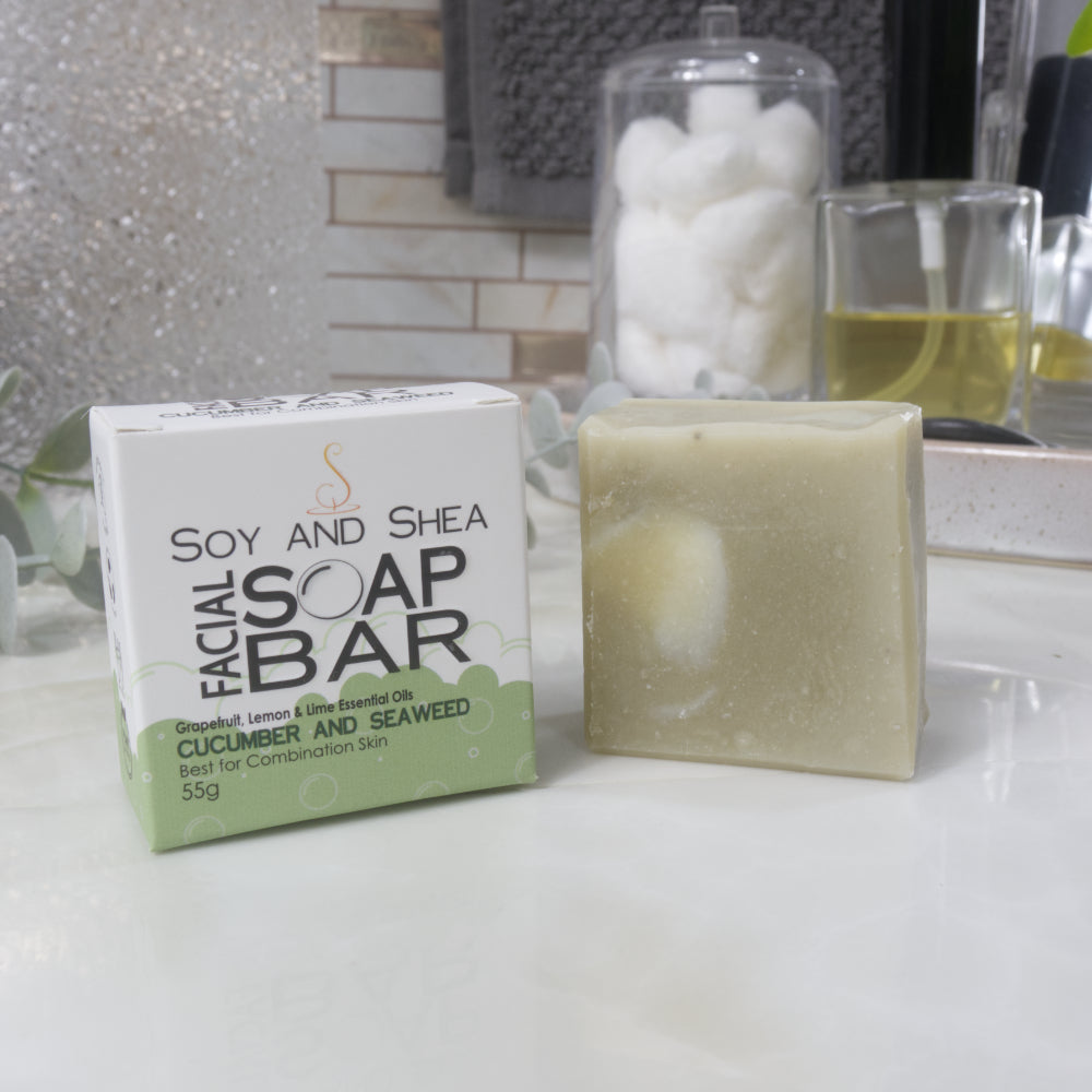 A pale green square bar of soap stands next to its packaging, a white and green box, on a bathroom countertop surrounded by accessories like perfume.  