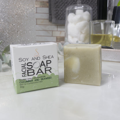 A pale green square bar of soap stands next to its packaging, a white and green box, on a bathroom countertop surrounded by accessories like perfume.  
