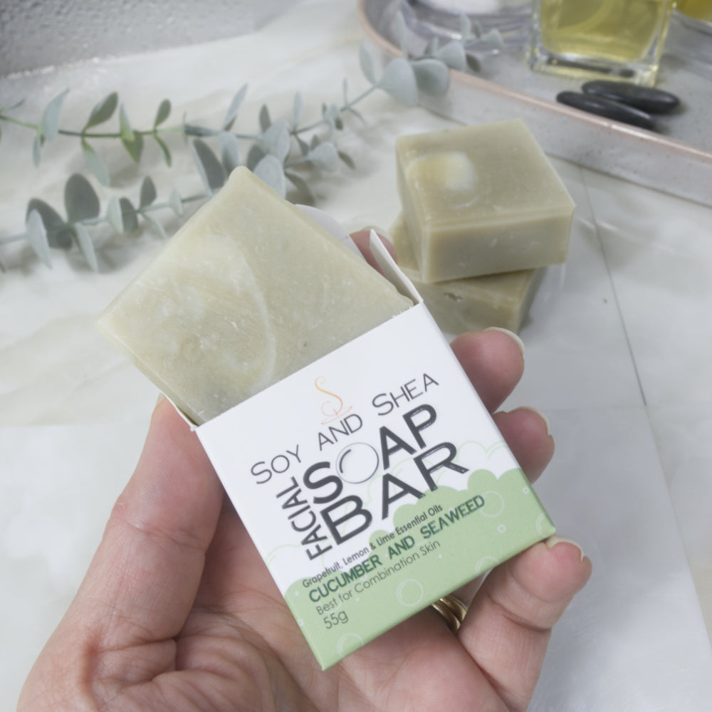 A hand holds a white and green box labelled as Facial Soap Bar with Cucumber and Seaweed.  The top of the box is open and a bar of pale green soap is shown coming out the top.  On the counter top is a stack of two more soaps along with some greenery