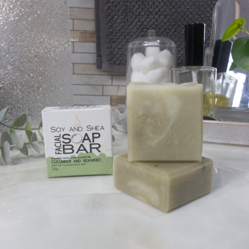 In the foreground a small square soap lays flat on a marble bathroom countertop with another standing upright on it.  Both soaps have a olive green colour with white swirls.  Behind stands a branded box announcing as Facial Soap Bar with Cucumber and Seaweed. 