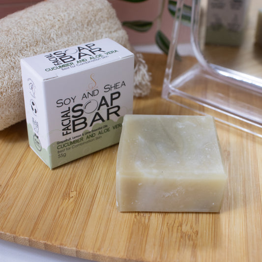 A small squarish soap bar in pale green lays flat on a wooden surface next to its packaging, a white box with a green border represent bubble foam.  In the background is a mirror, natural loofah and greenery.
