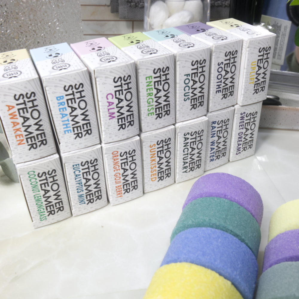 A set of shower steamer boxes in various scents arranged on a white surface, with several colorful shower steamer discs stacked in the foreground.