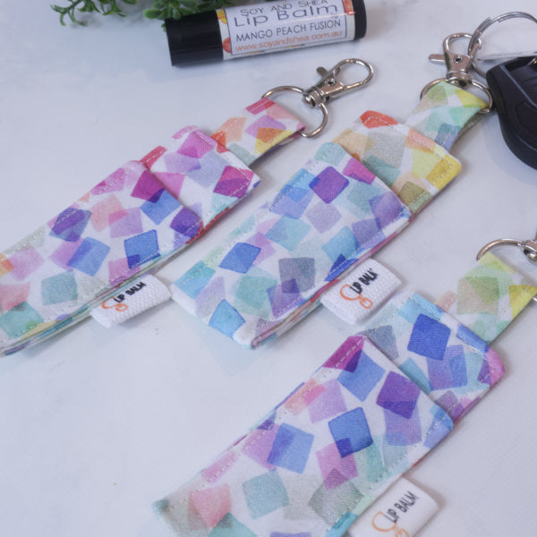 A selection of three clip balms showing variations in fabric design, lie on a wooden side with the middle Clip Balm attached to a car key.  A lip balm lays near-by along with some greenery.  The Clip balms have small watercolour squares in pastel rainbow colours scattered and overlayed over a white background.