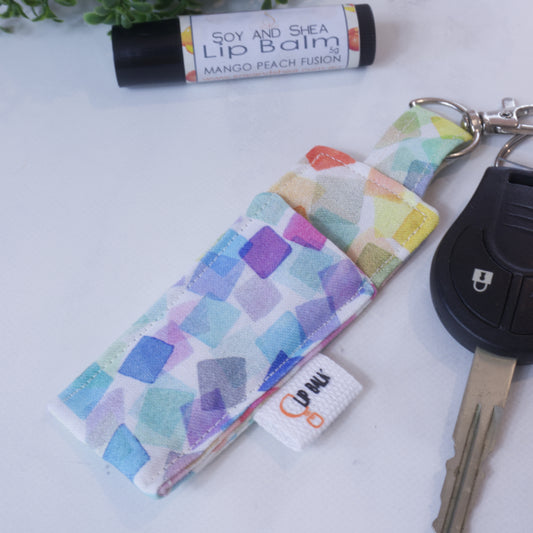 A lip balm holder lies on a wooden surface attached to a car key with a "lobster claw" clip.  To the side is a tube of lip balm surrounded by greenery.  The Clip Balm has rainbow coloured squares with a watercolour effect randomly placed over a white background
