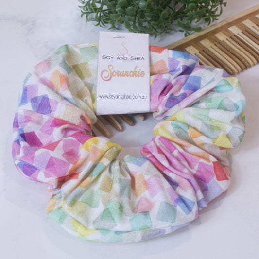 A colorful scrunchie sits on wooden surface resting against a wooden comb with greenery in the background.  A cardboard tag reads SOY AND SHEA SCRUNCHIE.    The scrunchie features an array of watercoloured squares in all shades of the rainbow scattered and overlayed across a white background