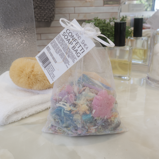 A clear organza bag with multicolored soap shavings visible inside, tied with a white ribbon with a swing tag reading CONFETTI SOAP BAG sits on a bathroom counter alongside a yellow bath sponge and various bottles.