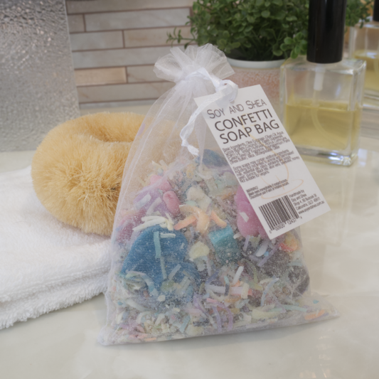 An organza bag filled with colour soap shreds and featuring a swing tag saying CONFETTI SOAP BAG, sits on a bathroom counter top with a body brush, perfume and potted plant.