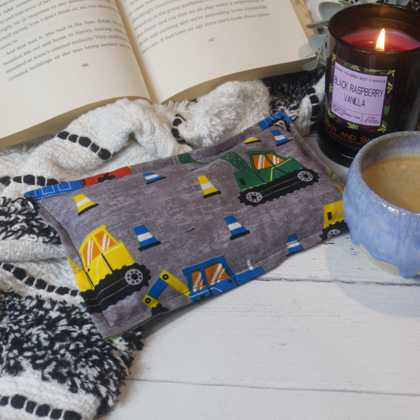 A mini rectangular heat pack rests against a black and white blanket and open book.  Next to it is a burning candle and filled coffee cup evoking a comfy scene.   The heat pack has a mottled grey background with a variety of construction vehicles and traffic cones in shades of blue, yellow and green
