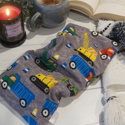 A small rectanglular Heat Pack rests on a wooden table top along with a black and white blanket, open book, burning candle and filled coffee cup evoking a cosy scene.  The Heat pack has has a grey mottled fabric covered in colourful construction vehicles and traffic cones in blues, green and yellow
