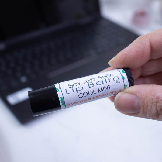 A hand holds a black tube of lip balm against a blurred background with an open laptop computer on a white surface.  The label has text LIP BALM COOL MINT on a white background with a border of green mint leaves.