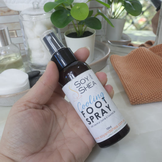 A hand holds a small amber bottle of Soy Shea Cooling Foot Spray in front of a bathroom counter with plants, jars, and a cloth.