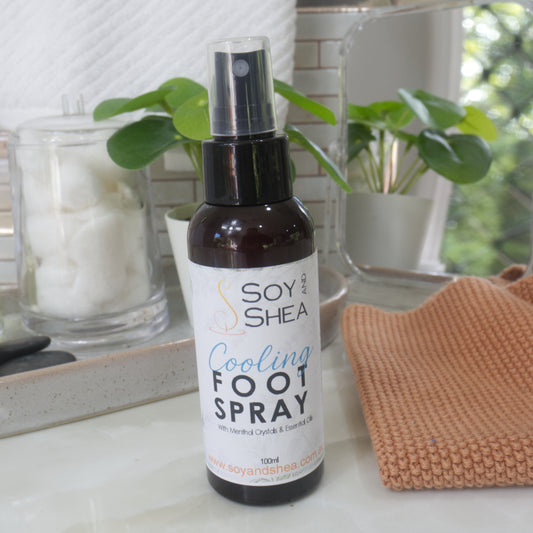 A bottle of Soy and Shea Cooling Foot Spray standing on a bathroom counter, with a mirror, potted plants, a jar of cotton pads, and a folded orange towel in the background.