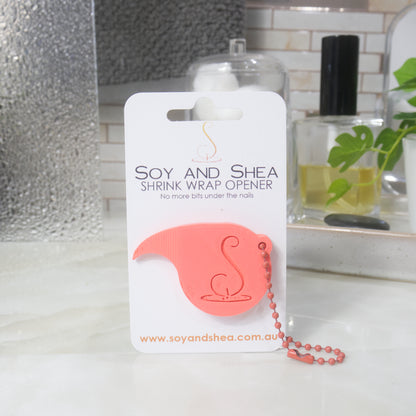 A Coral Shrinkwrap opener with matching ball and chain keyring is attached to a white card with product details.  The card is standing upright on a bathroom countertop with a bottle of perfume, a jar of cotton balls and towel in the background