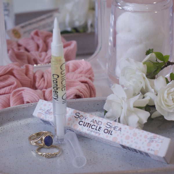 A clear, narrow tube with the lid removed showing a brush tip stands upright on a stone tray.  Beside it sits a cardboard box labelled CUTICLE OIL and a set of gold rings.  In the background is a bathroom counter top with a pink scrunchie, small mirror, jar of cotton balls and a some white  flowers.