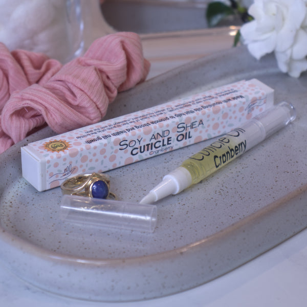 A clear, narrow tube with the lid removed showing a brush tip lies on a stone tray beside a cardboard box labelled CUTICLE OIL and a set of gold rings.  In the background is a bathroom counter top with a pink scrunchie, small mirror, and a some white  flowers.