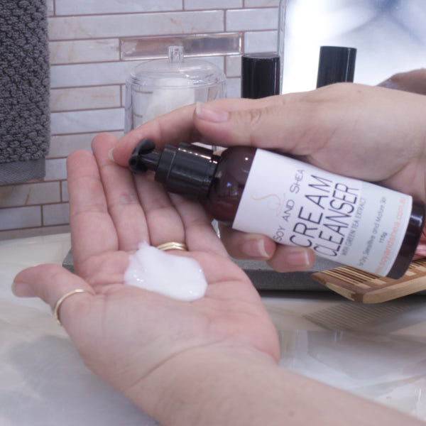 A person pumps Soy and Shea’s Cream Cleanser into their palm over a bathroom sink, with towels, jars, and bottles in the background.  The Cleanser is a white cream
