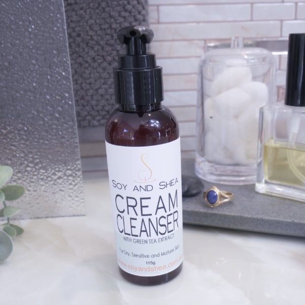 A pump bottle of cream cleanser on a bathroom counter, with a label that reads 'SOY & SHEA CREAM CLEANSER'. The background includes a glass jar with cotton balls and a ring on the counter.