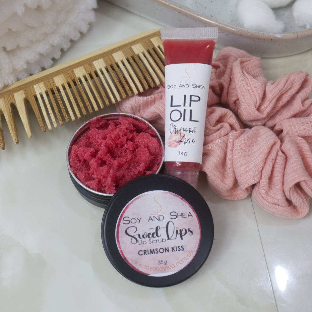 A small black tin with the lid removed revealing a red Lip Scrub sits on a marble tile surface next to a small, clear tube of red coloured Lip Oil.   This rests againsts a wooden comb and pink scrunchie. 