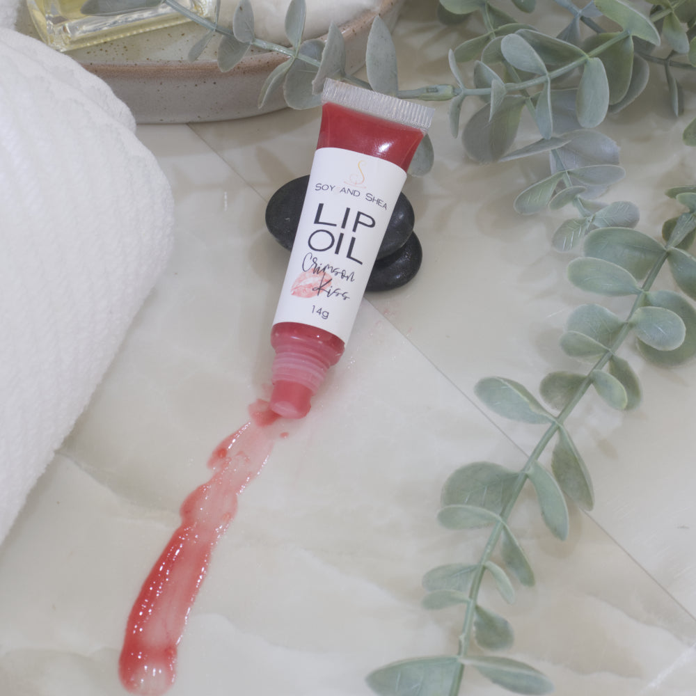A tube of Lip Oil lies on a marble tile surface next to a white towel and some eucalyptus leaves.   The tube is clear with a white product label and filled with a red Lip oil.  The cap is removed and a smear of Lip oil is on the bench showing the translucent effect it has