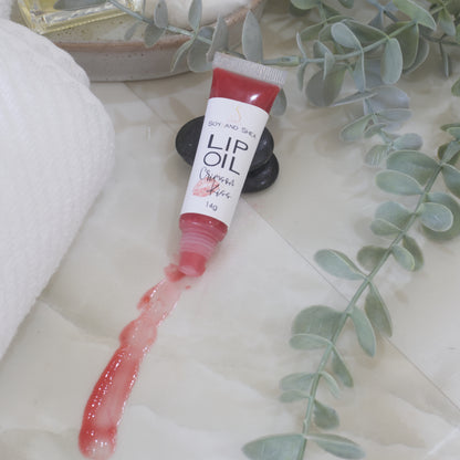 A tube of Lip Oil lies on a marble tile surface next to a white towel and some eucalyptus leaves.   The tube is clear with a white product label and filled with a red Lip oil.  The cap is removed and a smear of Lip oil is on the bench showing the translucent effect it has