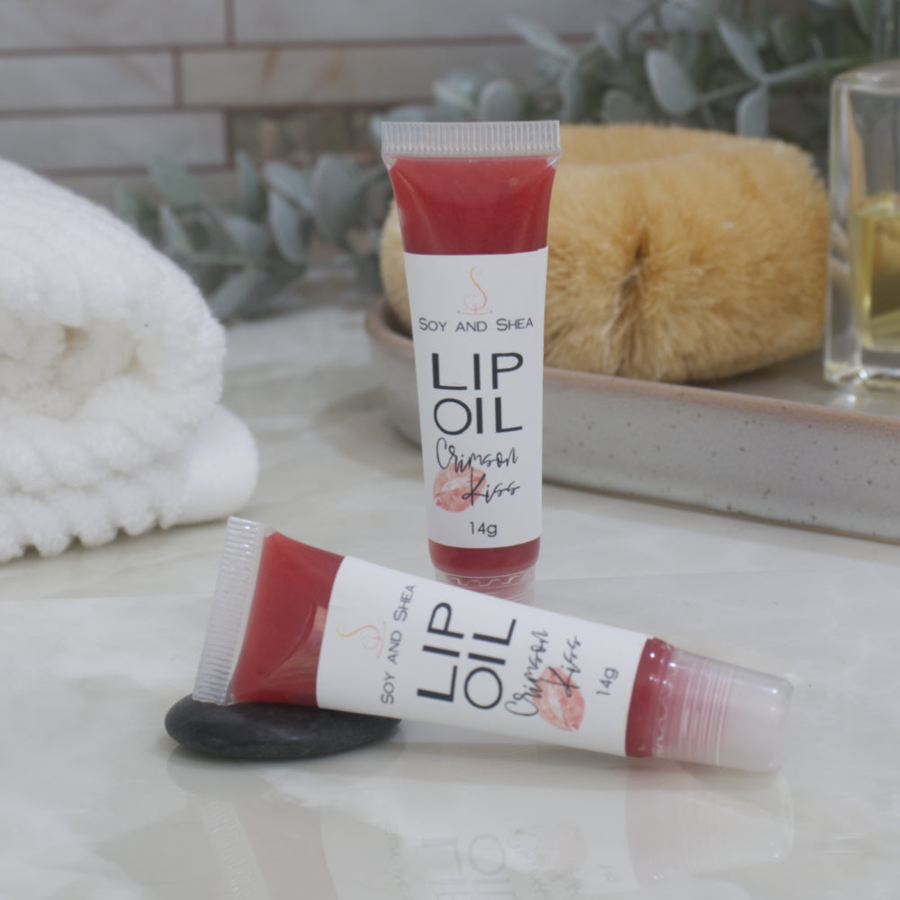 Two tubes of Lip Oil are presented on a bathroom countertop with a blurred background of towels, greenery and body brush.   One tube stands upright while the other lays flat in front of it.  The tubes are clear with a white product lable and are filled with a bright red Lip oil.