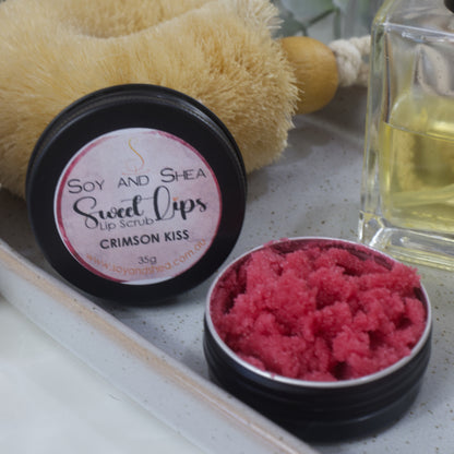 A round black tin sits on a stone tray along with a bottle of perfume and body brush.  The lid has been removed and is resting against the body brush revealing a red sugar scrub 