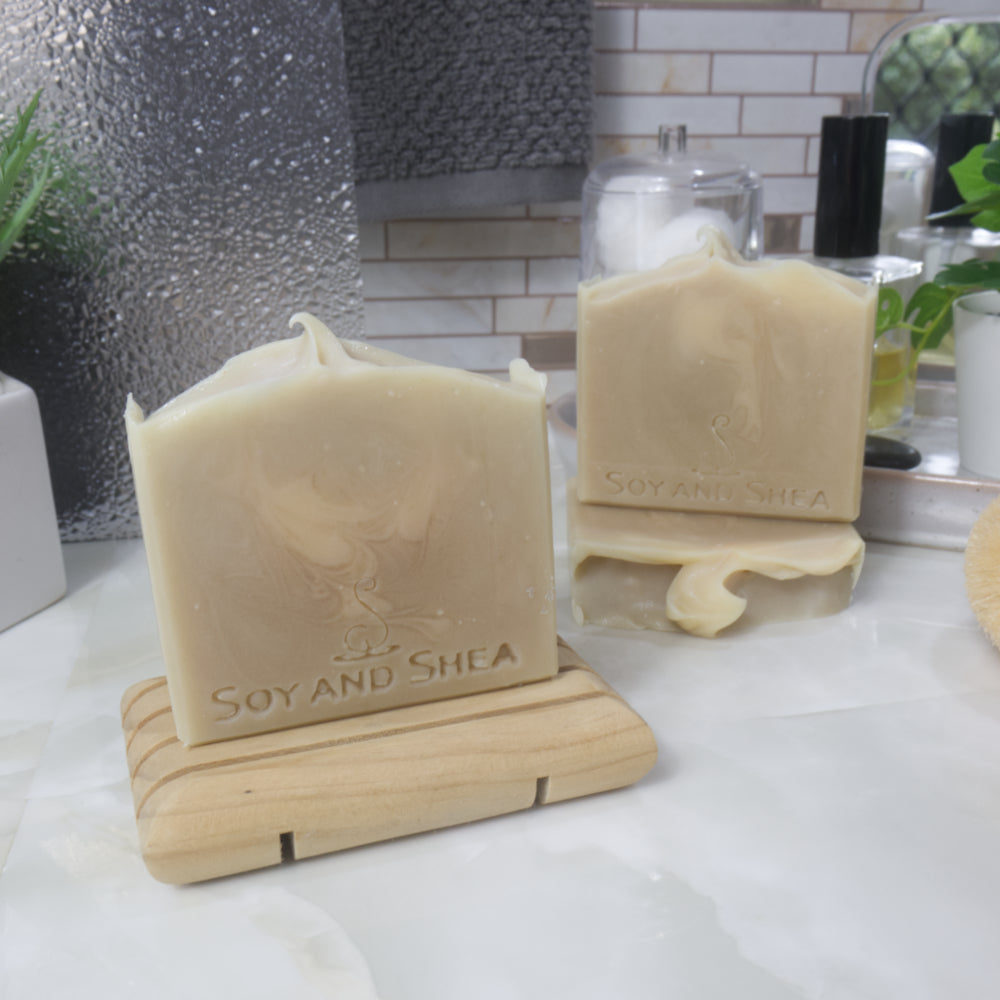 A selection of three soap bars are displayed on a bathroom countertop along with a bottle of perfume, a jar of cotton balls and a hanging towel.  One soap stands upright on a wooden soap dish towards the front left of the image.  behind that, a soap lays flay on the bench displaying a wavy soap top, with another soap standing on top.  The soaps are a cream-green colour with a pale cream swirl