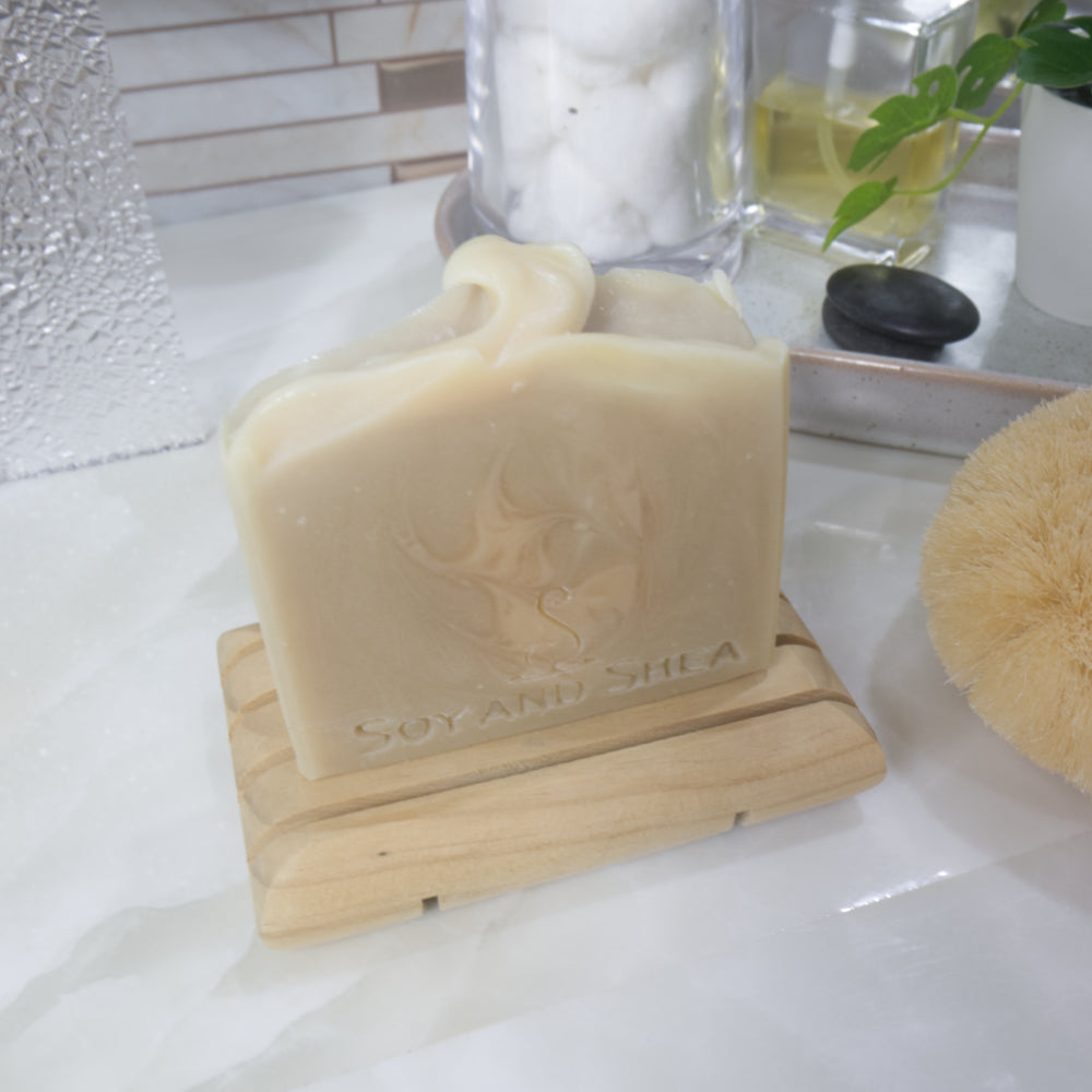 A bar creamy coloured soap with lighter cream swirls sits on a wooden soap dish on a bathroom countertop.  Behind is a stone tray with a bottle of perfume, a plant and a jar of cotton balls.    The image focuses on the wavy design on the soap top