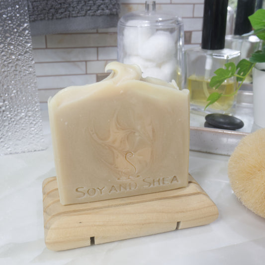 A bar creamy coloured soap with lighter cream swirls sits on a wooden soap dish on a bathroom countertop.  Behind is a stone tray with a bottle of perfume, a plant and a jar of cotton balls.  