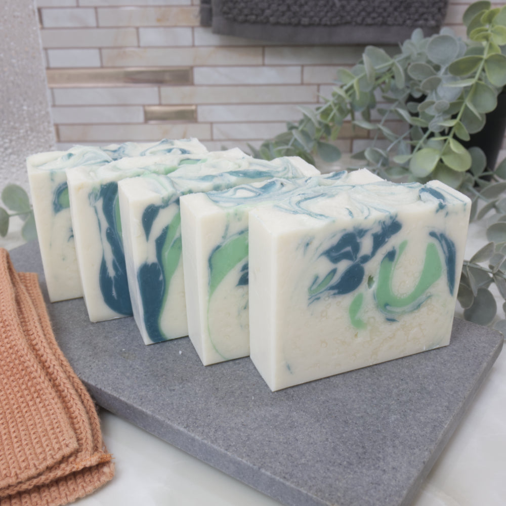 A row of fives soaps sit across a stone surface surrounded by green and bathroom items.   The soaps are white with light and dark green swirls.  Each soap has a different design