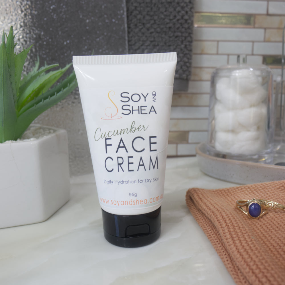 A white squeeze tube of Soy and Shea Cucumber Face Cream with a black cap, set on a marble countertop with a potted succulent, a glass jar of cotton pads, and a peach knit cloth in the background.