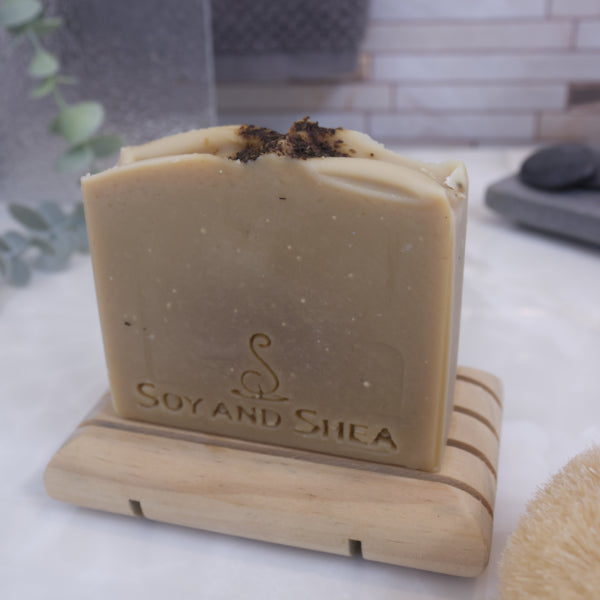 A pale khaki green sap bars stamped with SOY AND SHEA, sits on a wooden rack with a faded bathroom background.  Tea leaves are sprinkled on the top of the soap