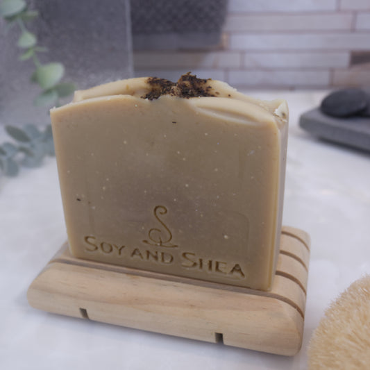A pale khaki green sap bars stamped with SOY AND SHEA, sits on a wooden rack with a faded bathroom background.  Tea leaves are sprinkled on the top of the soap