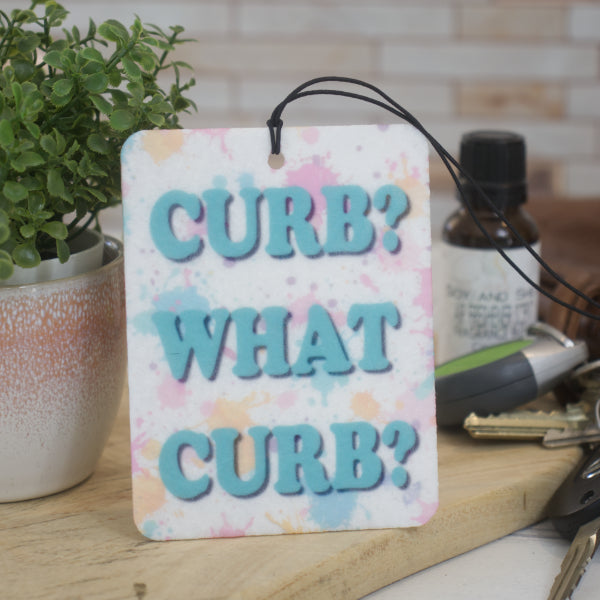 A car air freshener rests against a pot plant with the words CURB? WHAT CURB? in teal bold text ocer a paint splatter background.  Behind the air freshener is a bottle of scent and a set of keys.