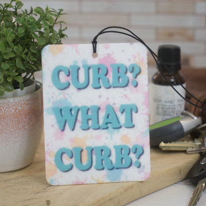 A car air freshener rests against a pot plant with the words CURB? WHAT CURB? in teal bold text ocer a paint splatter background.  Behind the air freshener is a bottle of scent and a set of keys.