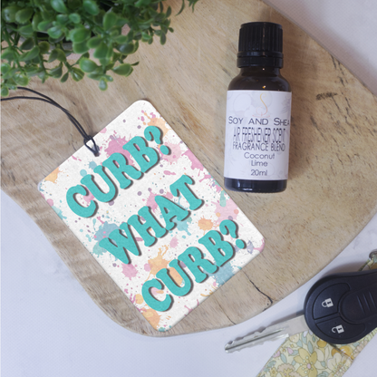 A car air freshener with the text 'CURB? WHAT CURB?' written in stylized letters on a multicolored background, lying on a wooden surface next to a bottle of scent
