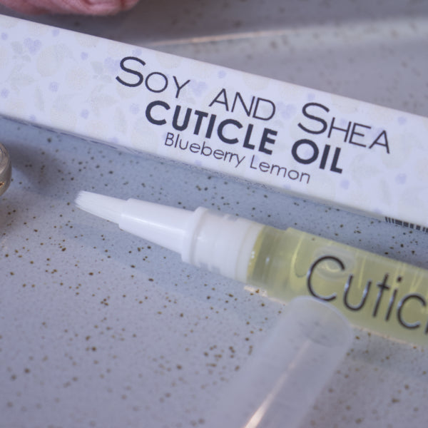 A close up image of the Cuticle oil pen showing the white brush tip. In the background is the box packaging with text CUTICLE OIL BLUEBERY LEMON and faded images of blueberries and lemons