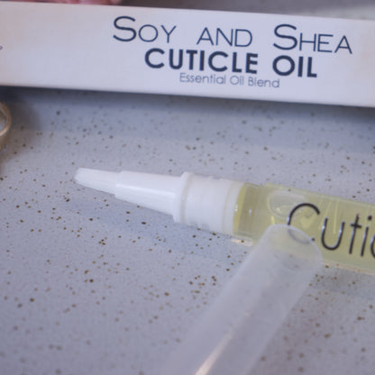 A close up image of the Cuticle oil pen showing the white brush tip. In the background is the box packaging with text CUTICLE OIL ESSENTIAL OIL BLEND