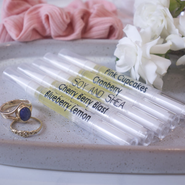 A set of cuticle oil in clear tubes with different flavors such as Pink Cupcakes,  Cherry Berry Blast, and Blueberry Lemon are displayed on a grey surface alongside a gold ring and a pink cloth with a white flower in the background.